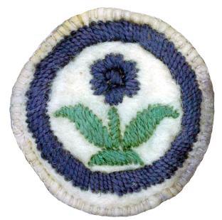 Image of a round felt badge with a logo of a flower embroidered in navy thread with green leaves, surrounded by a navy ring.