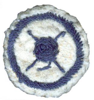 Image of round white felt badge with logo of ball of wool superimposed on crossed knitting needles embroidered in navy thread within an embroidered navy ring