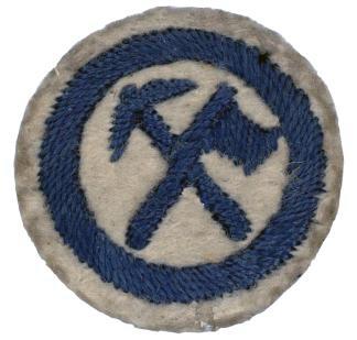 Image of a round white felt badge with a logo of a crossed axe and pick embroidered in navy thread, within a navy embroidered ring.